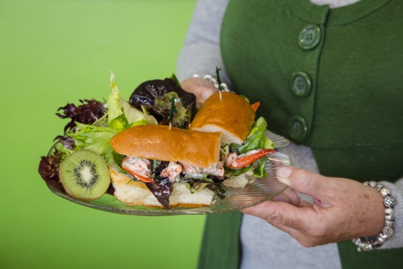 hands hold a lobster roll on a plate. there is a green background. 