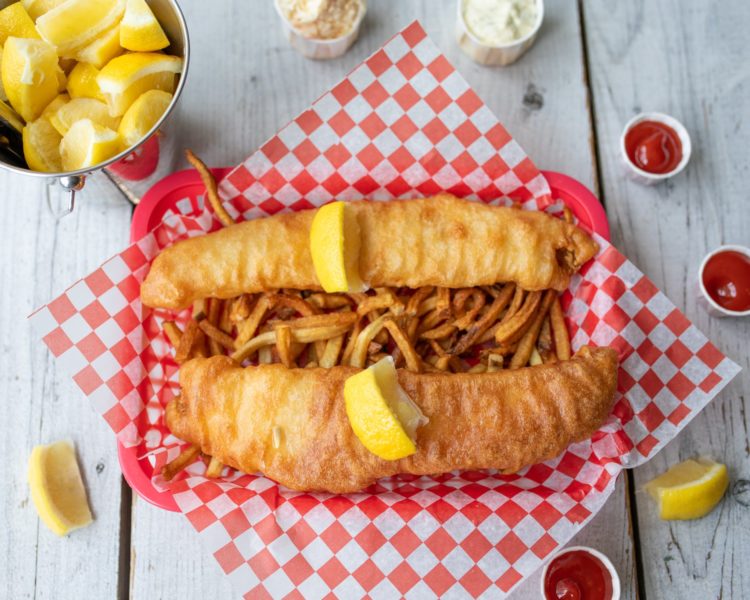 two pieces of fish and fries sit on a checkered paper in a red basket on a wooden table