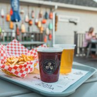 Two beers and fries sit on a tray on a picnic table. There are buoys on a wall in the background