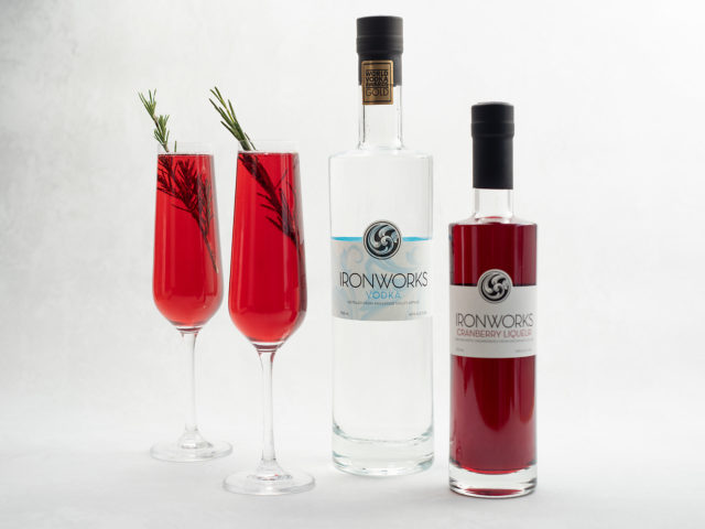 two red cocktails in champagne flutes sit by a bottle of Ironworks vodka and their cranberry liqueur