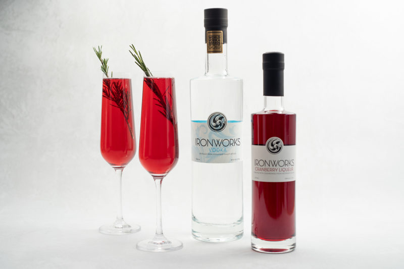 two red cocktails in champagne flutes sit by a bottle of Ironworks vodka and their cranberry liqueur