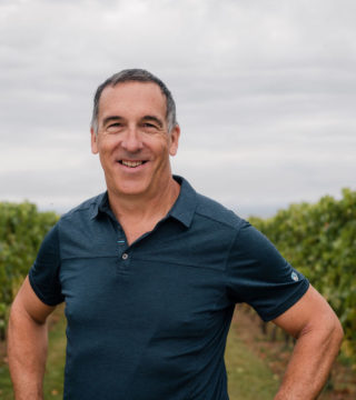 A photo of Hans Christian Jost. He is standing outside in a vineyard with a gray sky behind him.