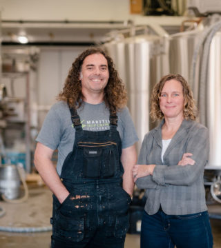 A photo of Boxing Rock's founding partners, Emily Cowin and Henry Pedro. They are standing in the brewery in front of silver brewing tanks.