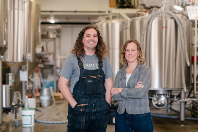 A photo of Boxing Rock's founding partners, Emily Cowin and Henry Pedro. They are standing in the brewery in front of silver brewing tanks.