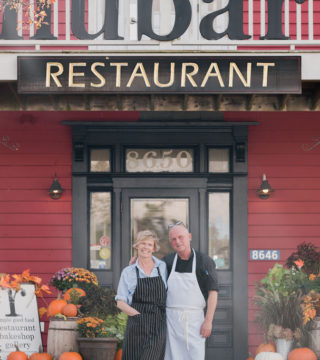 A photo of Rhubarb Restaurant owners and operators, Diane and Jim Buckle. They're standing on the stairs outside the restaurant, with the restaurant's logo visible above them.