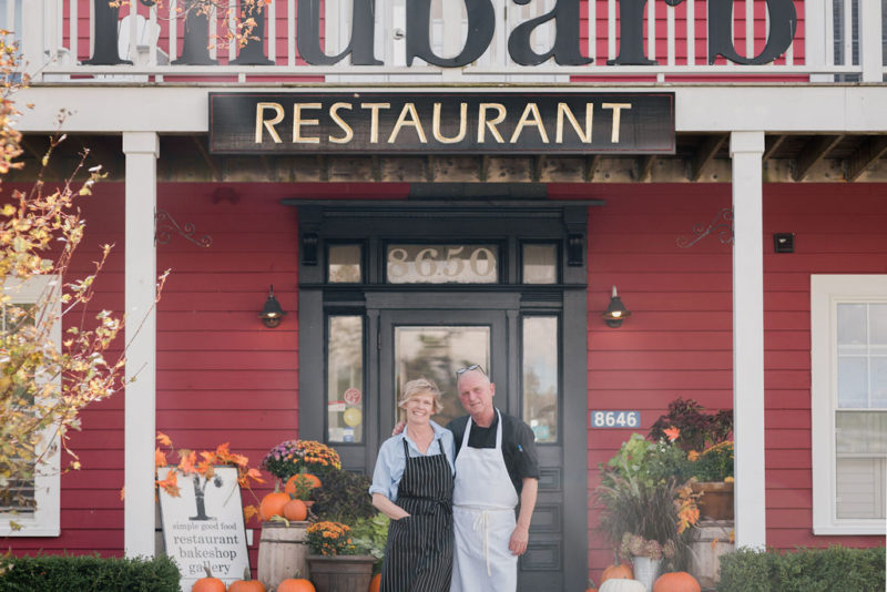 A photo of Rhubarb Restaurant owners and operators, Diane and Jim Buckle. They're standing on the stairs outside the restaurant, with the restaurant's logo visible above them.