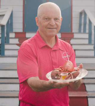 A photo of Shore Club owner Rhys Harnish standing on the stairs to the Shore Club. He is wearing a red shirt and holding a plate of lobster.