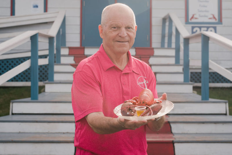 A photo of Shore Club owner Rhys Harnish standing on the stairs to the Shore Club. He is wearing a red shirt and holding a plate of lobster.