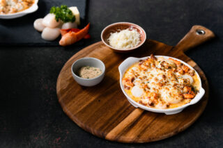 A dish of seafood gratin on a wooden serving board with a black background, surrounded by vegetables and two small bowls of ingredients.