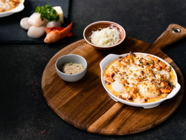 A dish of seafood gratin on a wooden serving board with a black background, surrounded by vegetables and two small bowls of ingredients.
