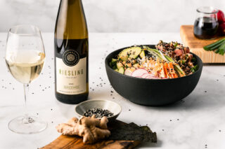 A tune poke bowl served in a black bowl on a marble counter. The bowl is surrounded by a glass and bottle of white wine, and a dish of sesame seeds and ginger on a wooden serving board.