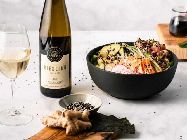 A tune poke bowl served in a black bowl on a marble counter. The bowl is surrounded by a glass and bottle of white wine, and a dish of sesame seeds and ginger on a wooden serving board.