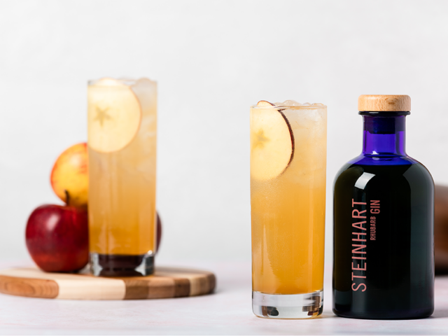Pictured are two orange-coloured cocktails, garnished with an apple slice. One glass is next to a blue bottle of Steinhart Gin, the other is on a wooden chopping board next to an apple. The background is white.