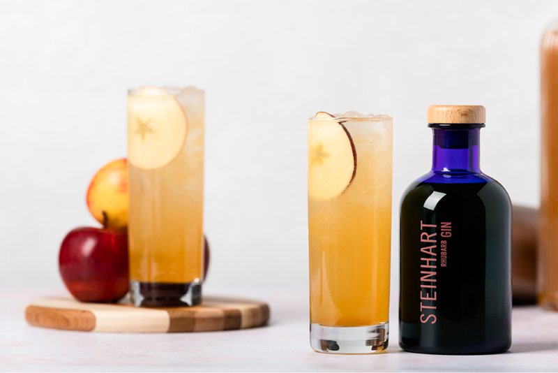 Pictured are two orange-coloured cocktails, garnished with an apple slice. One glass is next to a blue bottle of Steinhart Gin, the other is on a wooden chopping board next to an apple. The background is white.