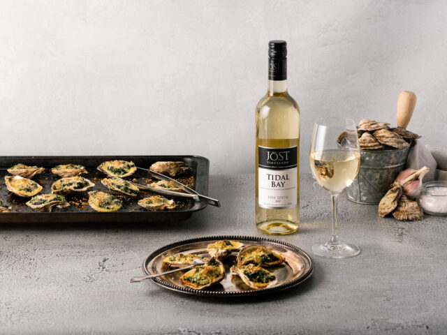 A plate of oysters Rockefeller is on a grey table with a baking sheet of oysters behind it. Also pictured is a glass and bottle of Jost Tidal Bay, and a bucket of fresh oysters and oyster shucker.