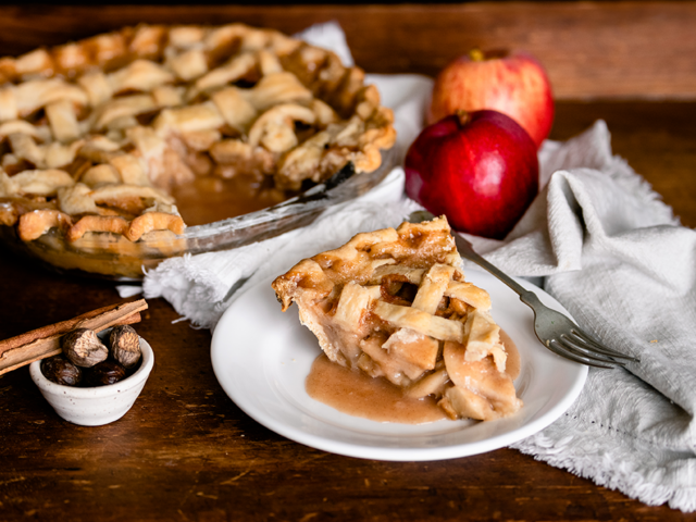 A piece of apple pie is on a white plate with the rest of the pie beside it. There are also two apples, and the background is brown wood.
