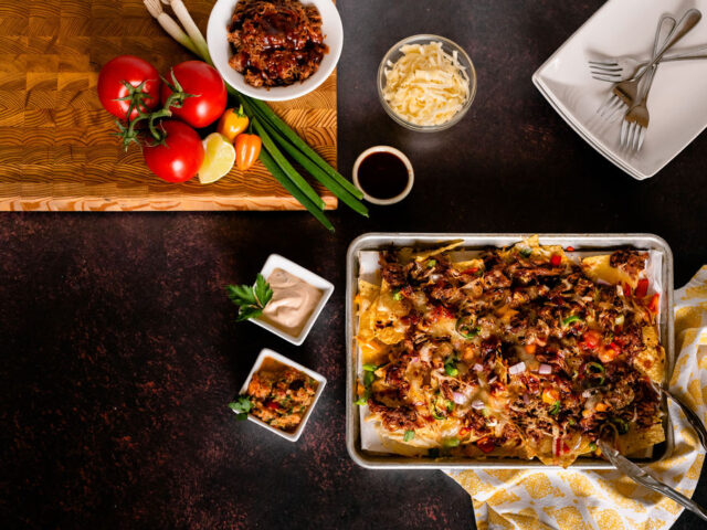 A tray of nachos on a dark background, surrounded by a yellow napkin, two ramekins of dip, and cutlery. Also pictured is a wooden chopping board with some produce on it.