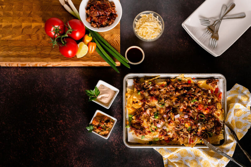 A tray of nachos on a dark background, surrounded by a yellow napkin, two ramekins of dip, and cutlery. Also pictured is a wooden chopping board with some produce on it.
