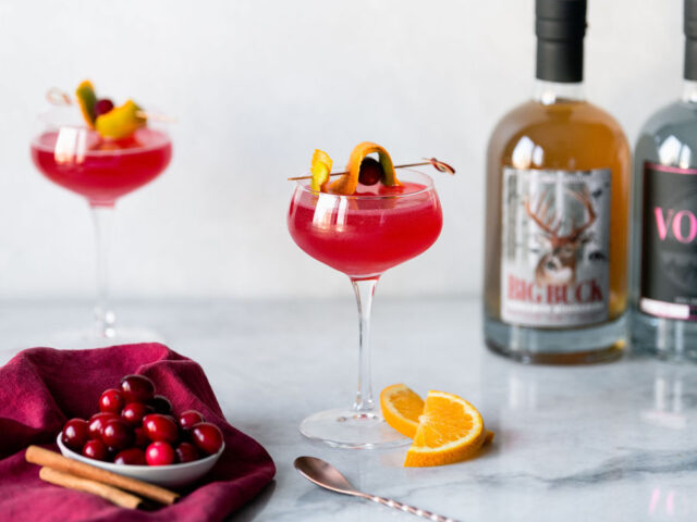 Two pink cocktails are in coupe glasses garnish with citrus rind in front of a white background. There are two orange slices next to the glasses, and one glass is on a red cloth napkin. Behind the glasses is a bottle of Still Fired vodka, and a bottle of Still Fired Moonshine.