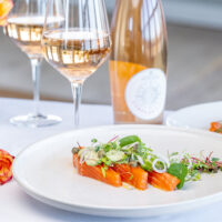 Trout on a white plate, garnished with greens. The plate is on a white table cloth surrounded by two glasses and a bottle of rosé, and some orange flowers.
