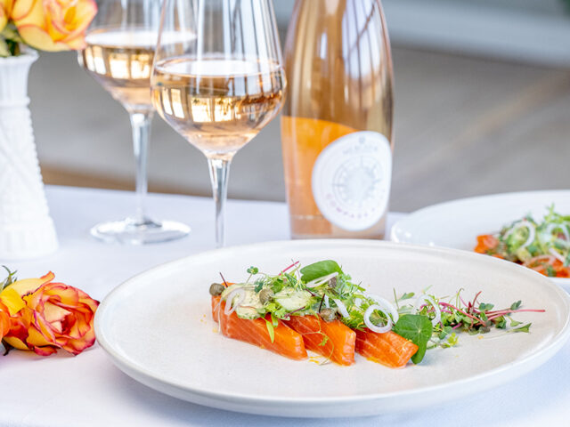 Trout on a white plate, garnished with greens. The plate is on a white table cloth surrounded by two glasses and a bottle of rosé, and some orange flowers.