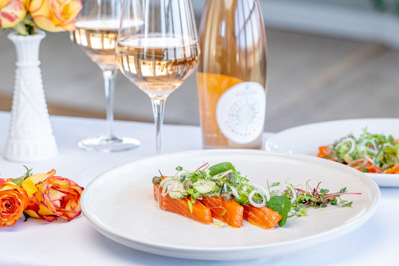 Trout on a white plate, garnished with greens. The plate is on a white table cloth surrounded by two glasses and a bottle of rosé, and some orange flowers.