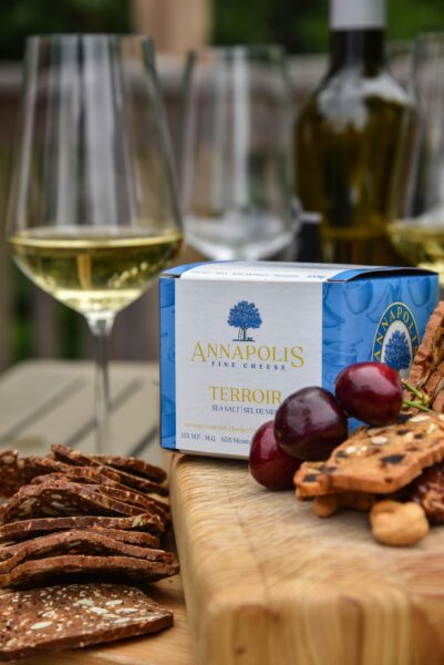 A photo of a box of Annapolis Fine Cheese's Sea Salt cheese sitting on a cutting board. There is a glass of white wine to the left, and grapes and crackers around the box.