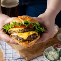 A photo of two hands holding a grass fed beef burger from Salt Shaker Deli. There are two patties, veggies and cheese.