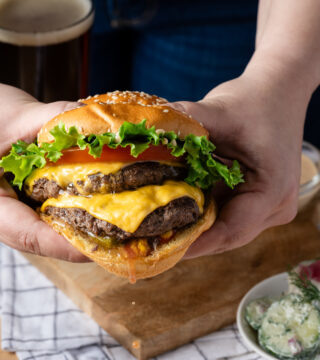 A photo of two hands holding a grass fed beef burger from Salt Shaker Deli. There are two patties, veggies and cheese.